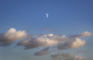 Half moon rising above fluffy clouds on a clear blue sky