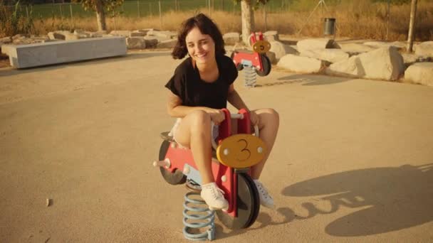 Брюнетка катается на качелях мотоцикле на детской площадке — стоковое видео