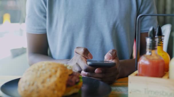 Primer plano irreconocible hombre negro mensajes de texto en el teléfono inteligente a la hora del almuerzo en la cafetería — Vídeo de stock