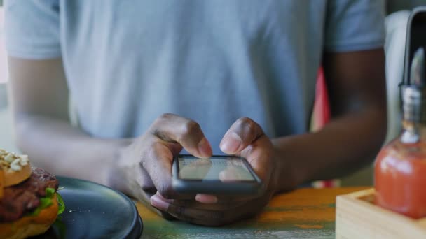 Primer plano irreconocible hombre negro mensajes de texto en el teléfono inteligente a la hora del almuerzo en la cafetería — Vídeo de stock