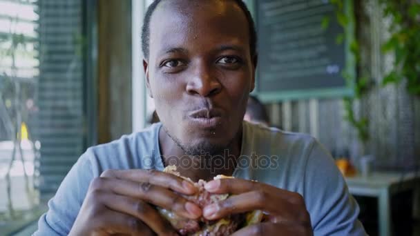 Счастливый черный парень смеется и ест вкусный мясной бургер — стоковое видео