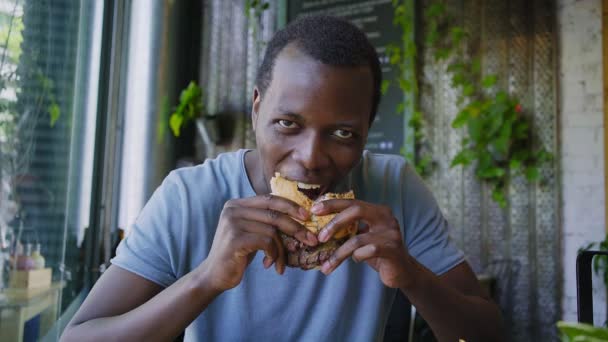 Glücklicher schwarzer Kerl lacht und isst leckeren Fleischburger — Stockvideo
