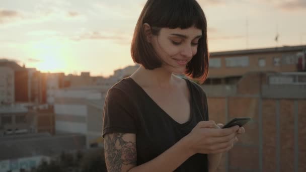 Aantrekkelijke brunette met gadget op dak bij zonsondergang — Stockvideo