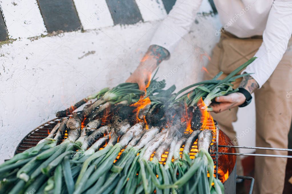 Man in white tshirt and arm tattoos makes barbecue on grill of spring onions and calcots, traditional spanish, catalan, mexican food