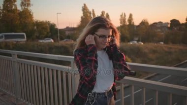 Ağır çekim trendy hipster kıyafeti sarı saçlı Genç kadının şehir sokak üzerinde yürümek, çıkar ve küçük kulaklık ile müzik dinlemek, Smartphone cep telefonu ile uygulama gezinmek, gülümseme ve dans