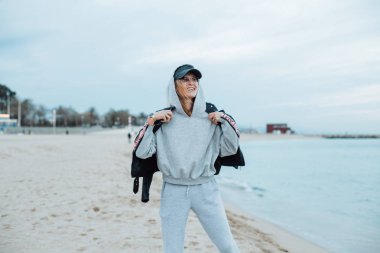 portrait of young woman in grey hoodie and sweatpants on beach clipart