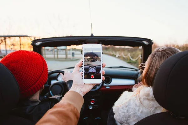 cropped view of hand taking photos on smartphone in cabriolet convertible car with friends
