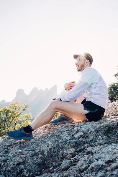 短いショート パンツや山や岩の上に座っているブロック ジャケット風を実行する強いと運動の若い男は 大規模なトレーニング セッションやトレーニング ウルトラ マラソンの後日没の風景を見下ろす — ストック写真