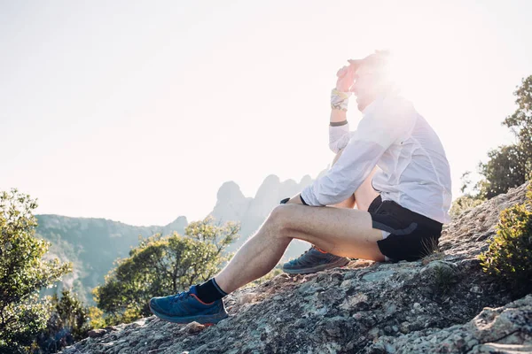 强壮和运动的年轻人在短的奔跑短裤和风阻拦夹克坐在山顶或岩石 俯瞰日落风景在广泛的锻炼会议或训练以后 超马拉松 — 图库照片