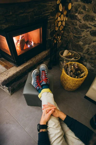 POV photo from above of man resting his feet in hard labor leather winter boots in front of warm and cosy hot fireplace, burning in living room or lobby of hotel in ski mountain resort