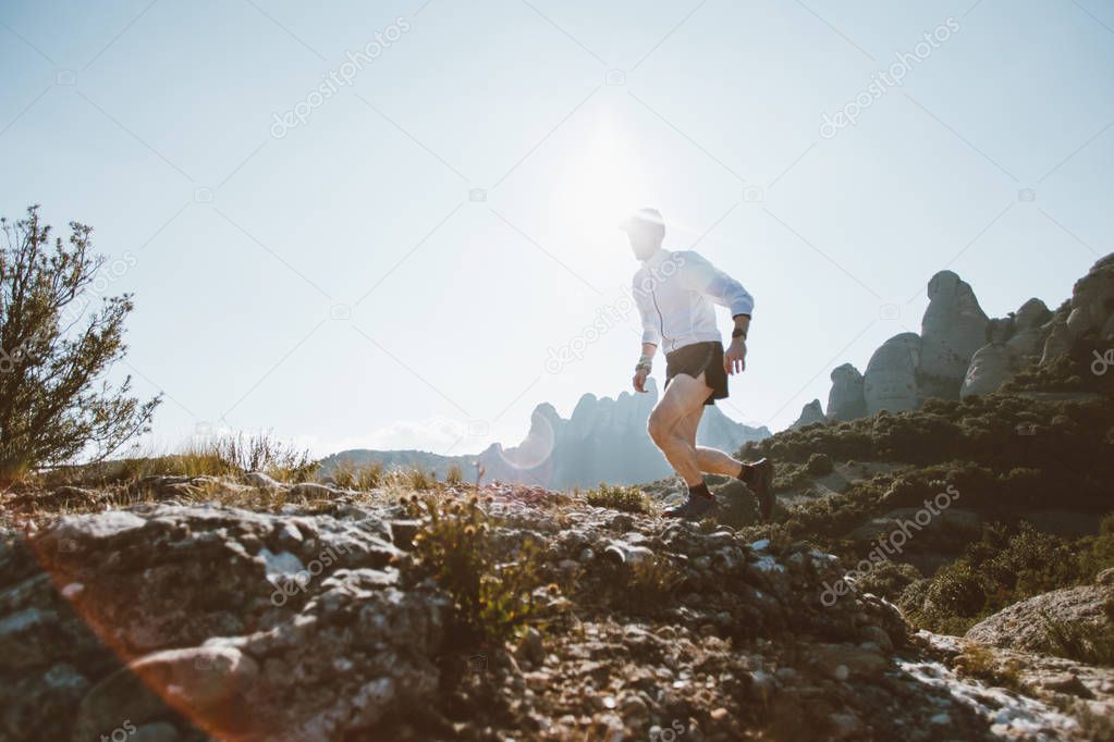 Professional trail runner, ultra distance athlete runs through rocky terrain high on mountain path or hiking trail, healthy lifestyle activity outdoors on sunny day. Active young man in sport wear