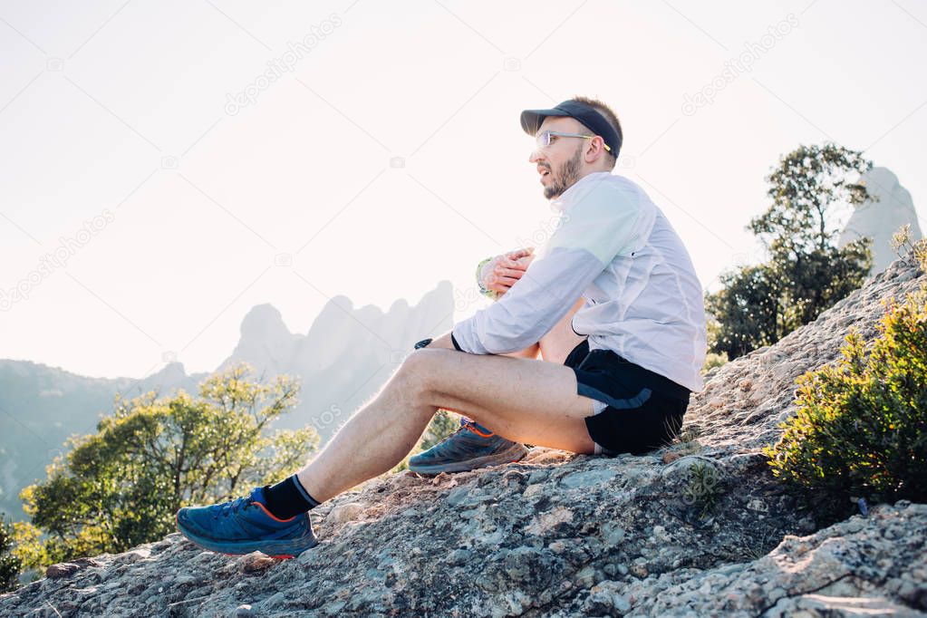 Sporty and strong athlete rests on top of mountain or cliff in sunset or sunrise light, with high mountains on background looks into distance overlooking path or trail, catching breath after workout run