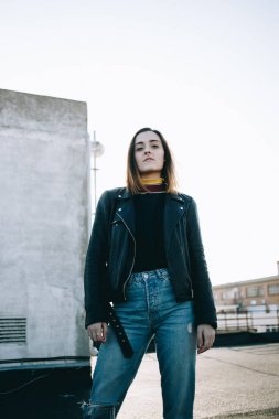 Fotoğraf makinesi güçlü güçlü bakışları ile serin ve korku veren genç güzel kadın deri ceket ve kot jeans, çatı modeli moda photoshoot için günbatımı teşkil etmektedir üzerinde duruyor bakar
