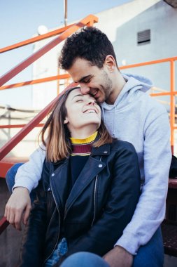Mutlu gülümseyerek ve trendy moda modern giysiler içinde genç çift gülüyor kırmızı yangın kaçış merdivenlerde otur, çatıda modeli photoshoot için poz. Serin ve kalça gençler veya millennials etkilendiği