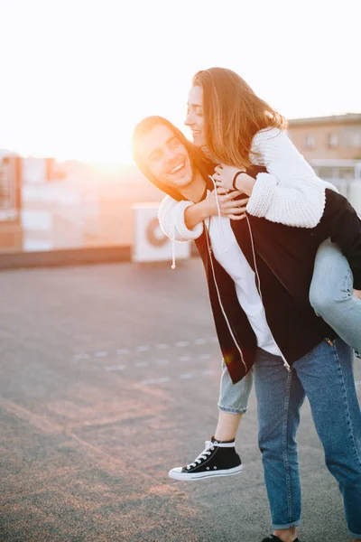 Happy and excited couple of hipsters laugh and have fun on rooftop in sunset. Young millennials in love, girl does piggy ride on back of boyfriend, honest feelings and true emotions of love