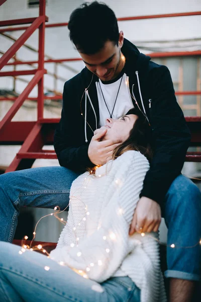 Tender and sensual, caring young couple during love story or wedding portrait photoshoot sit on fire escape staircase of new york style loft building. Cuddle and look at each other lovingly