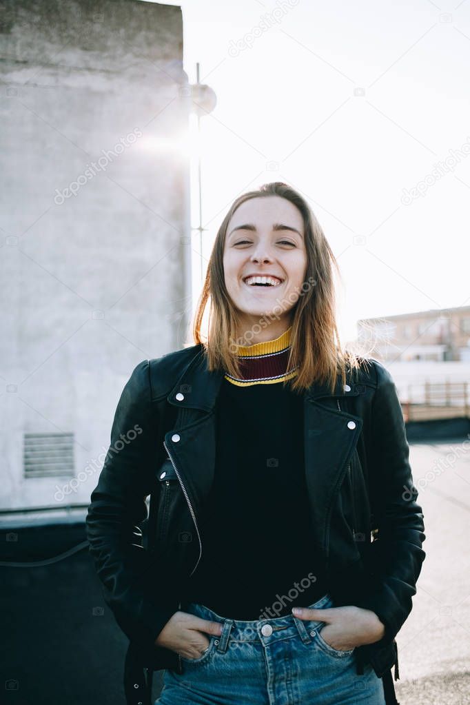 Beautiful and natural casual looking young woman laughs and smiles into camera, wears black leather jacket and turtleneck sweater, on sunset at rooftop, hipster and millennial lifestyle blogger