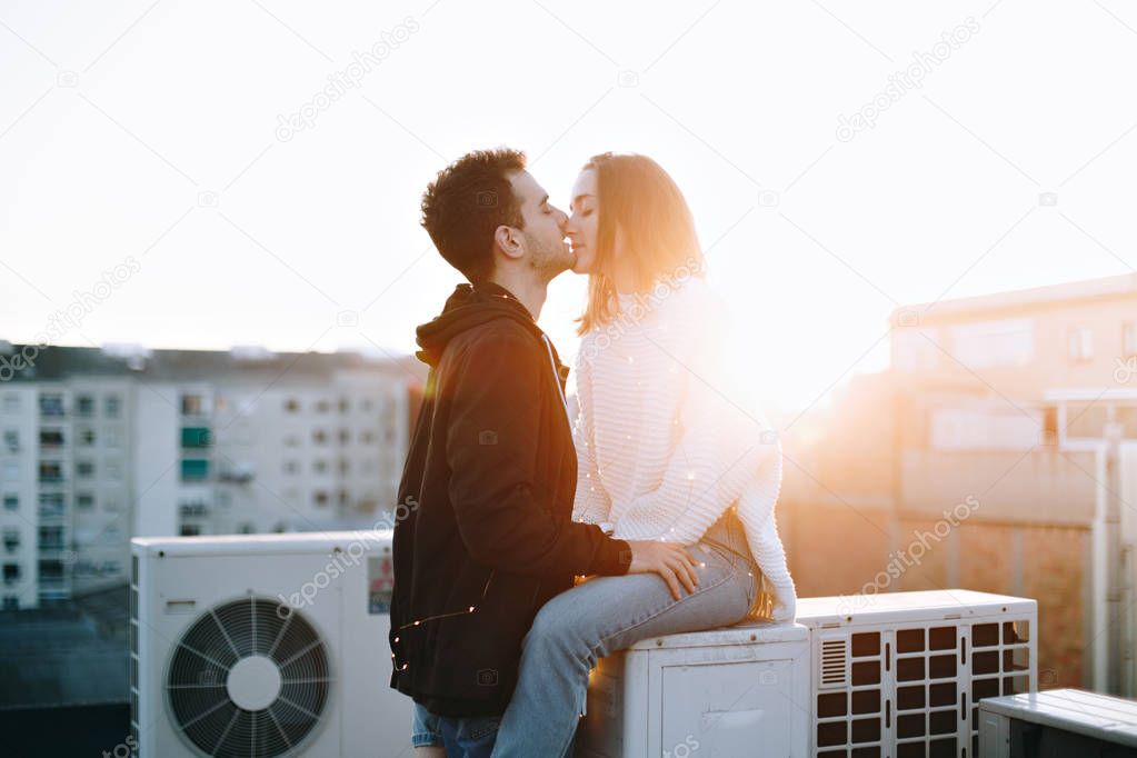 Cute romantic and tender sensual couple of young adults on date at rooftop on sunset in beautiful warm light rays, kiss and cuddle affectionately. Millennials in love or hipster blogger lifestyle