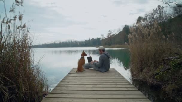 Freelancer travaille sur son ordinateur portable en plein air près du lac, assis sur l'étang — Video