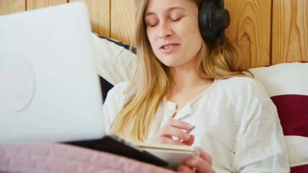 Fiatal nő fülhallgatóval és laptoppal az ágyban — Stock videók