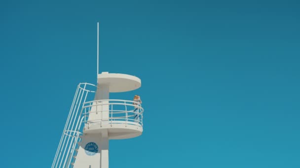 Κορίτσι στην παραλία ναυαγοσώστης πύργος σε απομονωμένο γαλάζιο ουρανό — Αρχείο Βίντεο