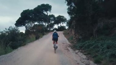 Formunda genç bir kadın orman yolunda çakıl bisikleti sürüyor.