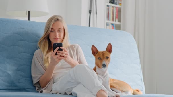 Jeune jolie blonde assise détendue sur canapé avec chien — Video