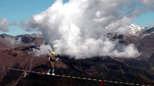 A girl in a yellow helmet walks on a suspension bridge amid beautiful mountains with clouds — Stock Video