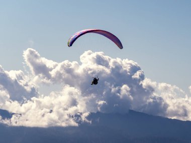 Mavi gökyüzüne ve açık havada beyaz bulutlara karşı dağların üzerinden uçan paraglider.