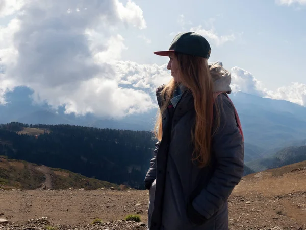 Ένα κορίτσι σε ένα καπέλο και ένα γκρι σακάκι κοιτάζει στην απόσταση με φόντο ένα βουνό κοιλάδα του μπλε ουρανού και τα σύννεφα — Φωτογραφία Αρχείου