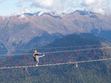 Bir kız uçurumun kenarındaki asma köprüde duruyor. Tırabzanlara tutunuyor ve ayağını karlı dağ zirvelerinin arka planına doğru kaldırıyor.