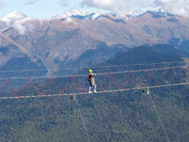 Bir kız uçurumun kenarındaki asma köprüde bir dağ sırasının arka planında yürüyor.