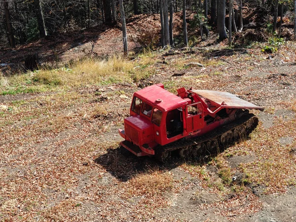 Sebuah traktor merah berdiri di tanah berkerikil abu-abu di antara dedaunan kering dengan latar belakang hutan — Stok Foto