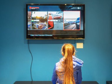 Sochi, Russia - 13 October 2019. A girl with long golden hair plays a game console looking at the screen hanging on a blue wall clipart