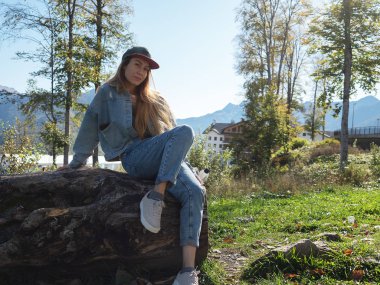 Şapkalı ve kot pantolonlu bir kız yeşil çimenlikteki büyük bir taşın üzerinde oturuyor.