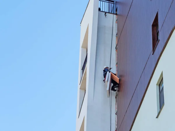 An industrial climber hanging from ropes restores a fragment of a building wall