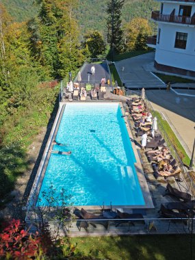 Sochi, Russia - 13 October 2019. People sunbathe on sun loungers and swim in the pool with clear blue water in the open air among the mountains and trees.