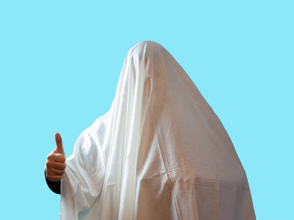 A man covered with a white sheet like a ghost shows a thumb on a blue background