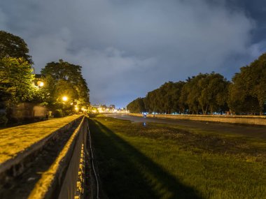 Kaldırımdan şehir nehrine kadar gece manzarası çimlerle, ağaçlarla ve parlak ışıklarla kaplıydı.
