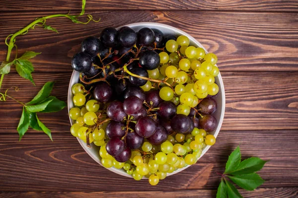 Ovanifrån skål med olika druvor: röda, vita och svarta bär och några gröna blad på den mörka trä bakgrunden. Selektivt fokus. — Stockfoto