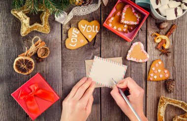 Üstten Görünüm erkek yazma isteklerine bir Noel tebrik kartı üzerinde eller. Ahşap masa dekor, ev yapımı kurabiye kalp şeklinde kutusuyla ile. Seçici odak. Festival Hediyelik.