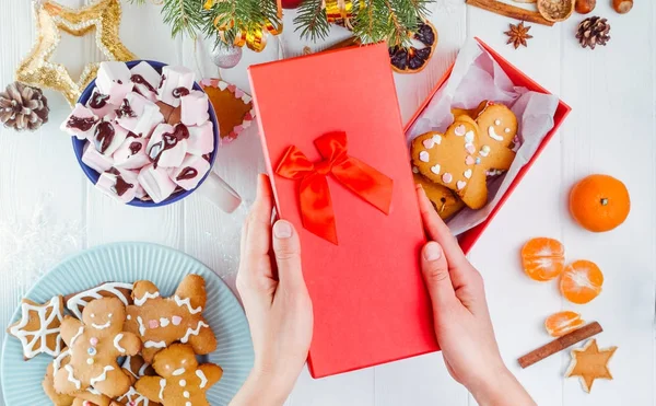 Üstten Görünüm kadın el yapımı geleneksel Noel zencefilli kurabiye adam kurabiye ile kırmızı hediye kutusundan kapak tutan eller. Beyaz ahşap masa dekorasyonu ile. Noel kavramı. Seçici odak.