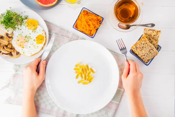 Top view female hands holding knife and fork and white plate with vitamin pills on the served wooden table with breakfast meal. Pill instead of food. Healthy diet concept. Selective focus. — Stock Photo, Image
