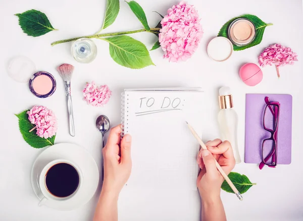 Las manos femeninas escriben Para hacer una lista en el lugar de trabajo blanco con accesorios femeninos, taza de café, cuaderno, vasos y flores de glicina. Fondo de moda, concepto de blog de belleza . —  Fotos de Stock