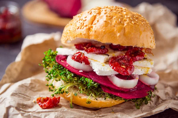 Hamburguesa vegetariana con rodajas de remolacha, brotes microverdes, queso tofu, tomates secos y cebolla en papel kraft sobre fondo de piedra oscura con inhrediantes. Enfoque selectivo suave. Copiar espacio . — Foto de Stock