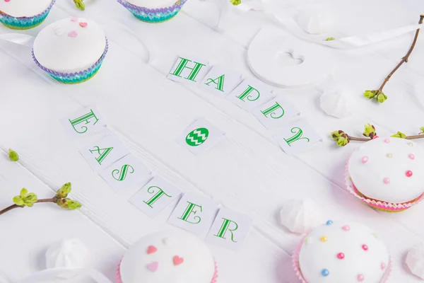 Feiertagskomposition aus fröhlichen Osteraufschriften, Zweigen mit jungen grünen Trieben, verzierten Cupcakes, Merengue-Bonbons, Vogelfigur auf Holzgrund. Kunstkonzept. Selektiver Fokus. — Stockfoto