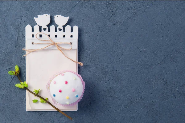 Simple composición de vacaciones de Pascua de cupcake decorado, rama con brotes jóvenes de vegetación y cuaderno de artesanía con vacío en blanco sobre fondo de piedra oscura. Enfoque selectivo. Espacio para texto . — Foto de Stock