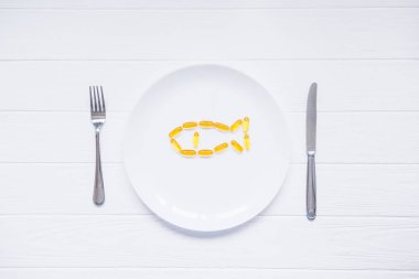 Üstten Görünüm morina karaciğer yağı kapsülleri, Omega 3, Vitamin D beyaz yuvarlak plaka üzerinde balık şeklinde bıçak ve çatal Beyaz ahşap masa üzerinde hizmet verdi. Seçici odak, metin için yer.