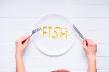 Morina karaciğer yağı kapsülleri, Omega 3 beyaz tahta masada tarafından bıçak ve çatal yuvarlak tabak balık kelime yazı ile altında tutarak üstten görünüm eller. Sağlıklı yaşam. Seçici odak, metin için yer.