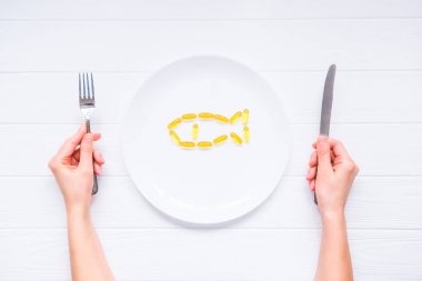 Üstten Görünüm bıçak ve çatal morina karaciğer yağı kapsülleri ile yuvarlak plaka altında tutan eller, Omega 3, D vitamini balık beyaz tahta masada şekil kadın. Seçici odak, metin için yer.